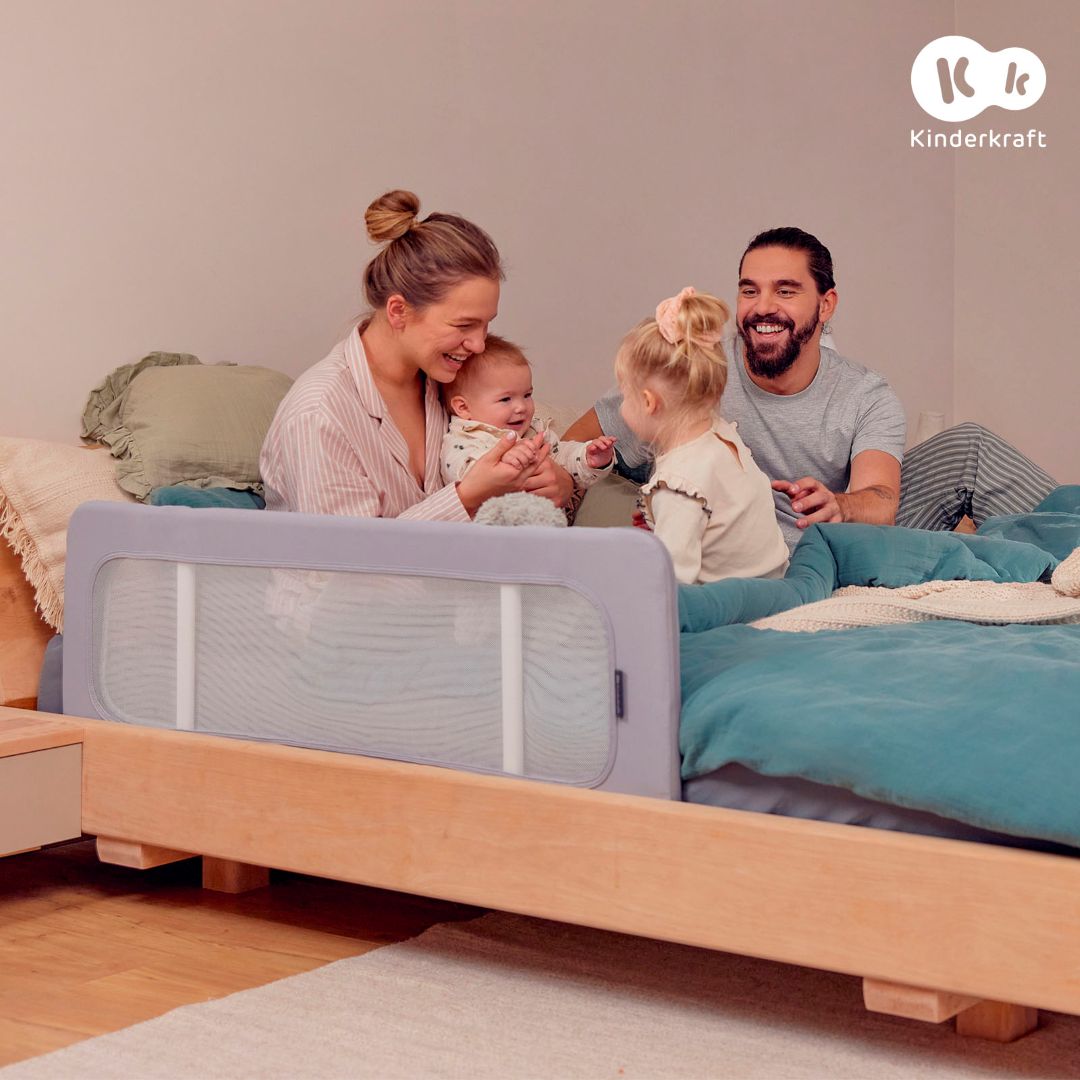 Familia con bebé y niña pequeña en cama protegida con barrera Kinderkraft GUARD & DREAM gris, segura para niños de 18 meses a 5 años, universal para cualquier colchón