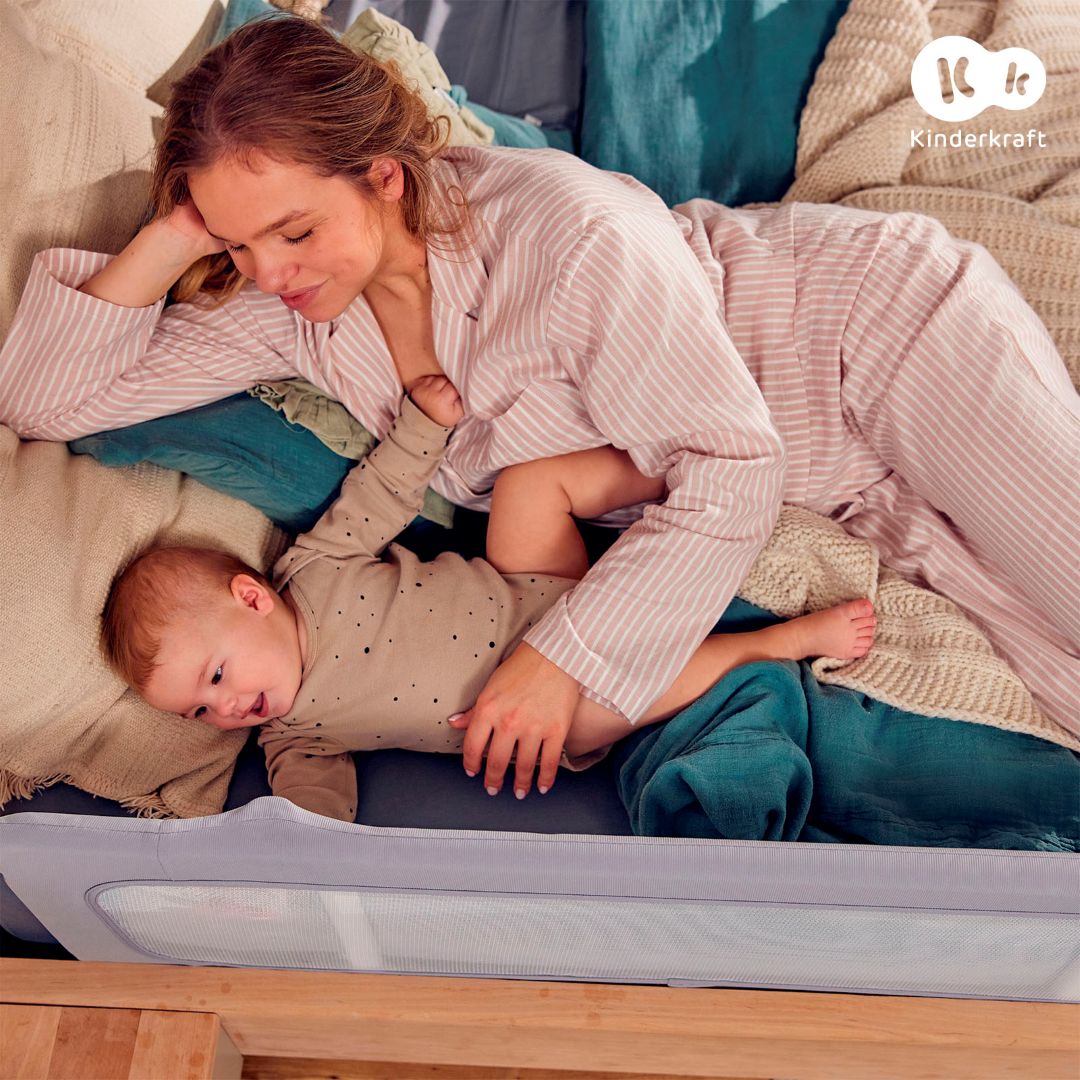 Mamá junto a su bebé en cama protegida con barrera Kinderkraft GUARD & DREAM gris, bordes acolchados suaves y malla transpirable para dormir seguros