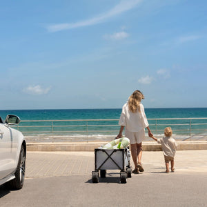 Mamá arrastrando el carrito Kinderkraft ROLLSTER cargado con accesorios de playa junto a su hijo, ruedas todoterreno 360° para cualquier superficie