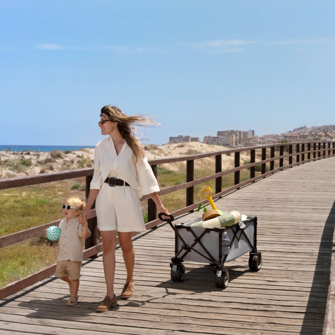 Mamá tirando el carrito Kinderkraft ROLLSTER por pasarela de madera junto al mar con accesorios de playa, ligero y manejable para paseos en familia